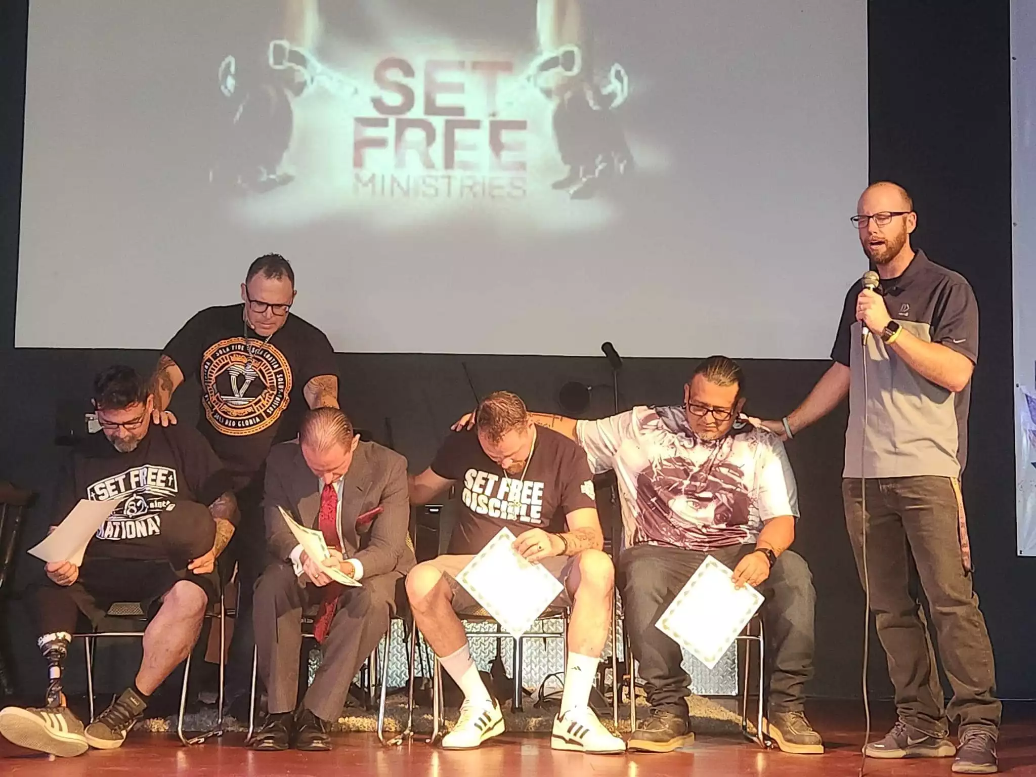 Men being prayed over on stage at a Set Free Ministries service while a leader speaks into a microphone