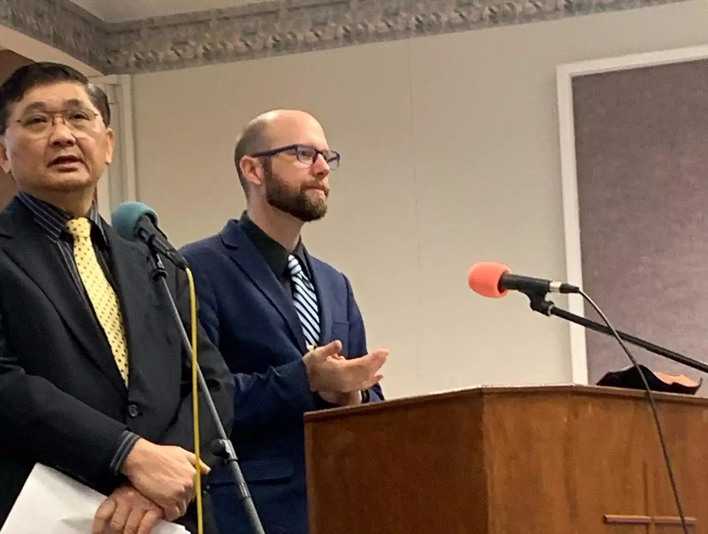Two men speaking at a wooden podium with microphones during an association gathering
