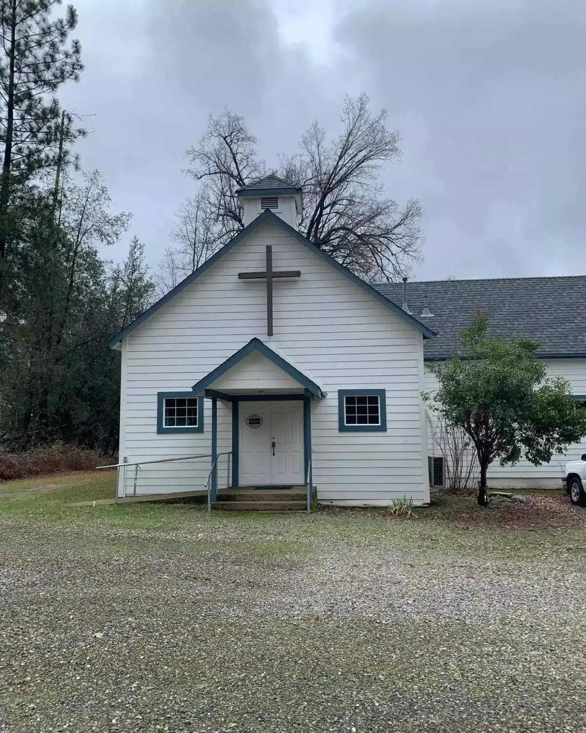 A small white clapboard church with a wooden cross above the entrance, on a gravel lot among pine trees